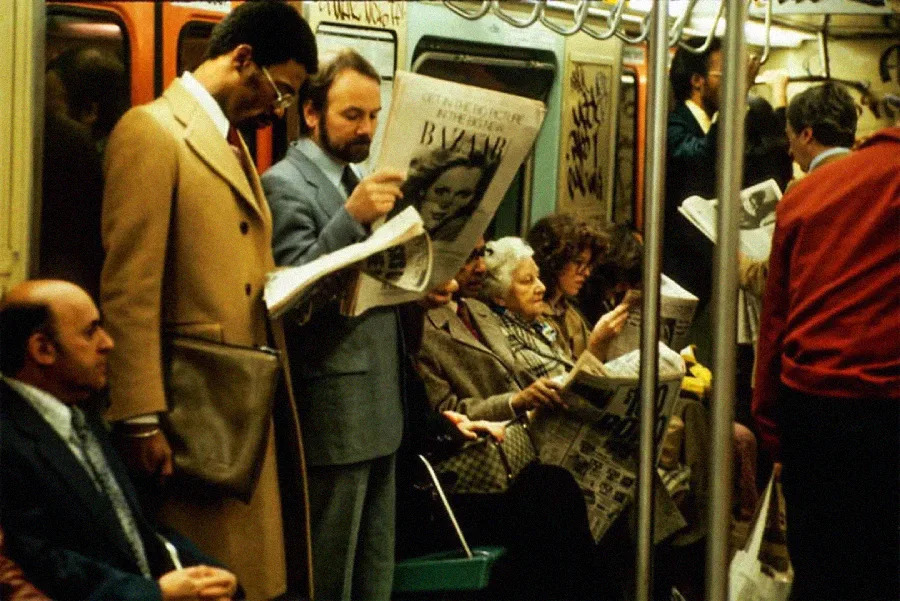 A crowded subway car with people sitting and standing, many reading newspapers. Some wear coats and formal attire, and graffiti is visible on the walls of the train. The scene appears to be from past decades.