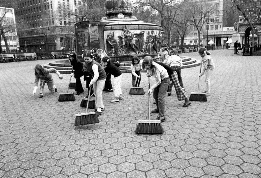A group of children sweeping a hexagonal-tiled plaza near a large, ornate fountain in an urban park on a cloudy day. Buildings and leafless trees are visible in the background.