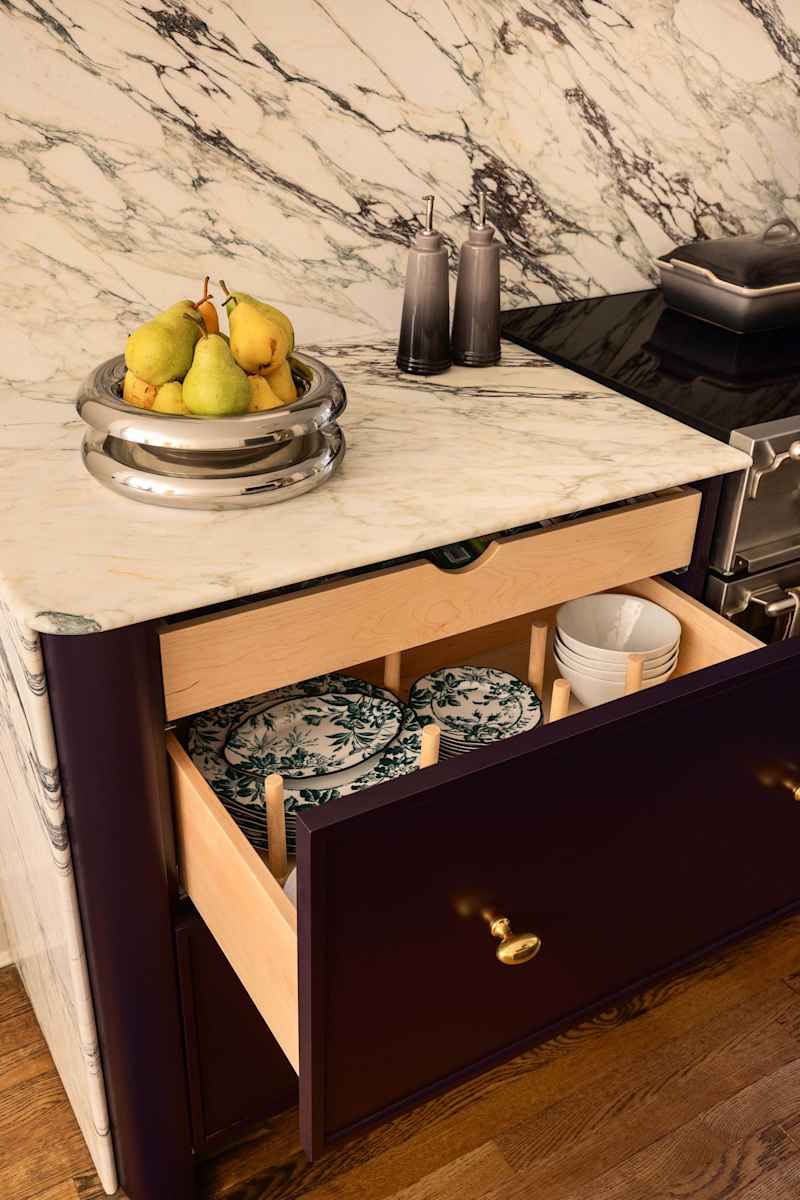 A modern kitchen countertop with a marble backsplash, a bowl of pears, and an open drawer displaying decorative plates and bowls.