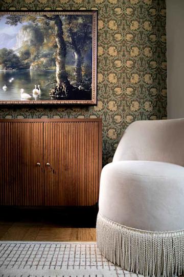 A cozy corner featuring a plush white chair with fringe, a wooden cabinet with ribbed texture, and a framed landscape painting.