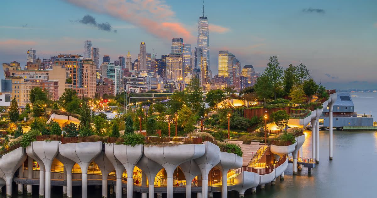 A scenic photo of New York City's floating island, "Little Island". The island is well lit and there is a beautiful city skyline in the distance.
