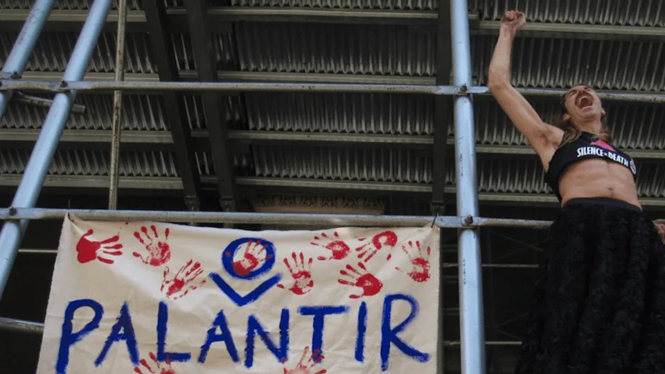 On the scaffolding of a building, a person raises their fist and yells. Beside them, a banner reads "Palantir" with several bloody handprints around it.