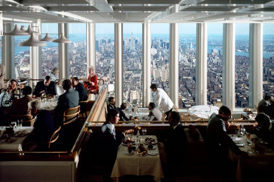 Elegant restaurant diners sit at tables near large windows offering a panoramic daytime view of New York City's skyline and skyscrapers. Waitstaff attend to guests, and sunlight brightens the modern, sophisticated interior.