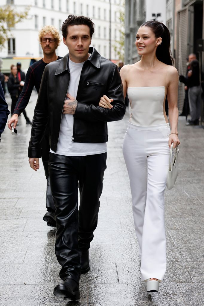 Brooklyn Beckham wears white t-shirt, black leather jacket, matching pants, black boots, and Nicola Peltz wears white jumpsuit with sheer details walking in paris street