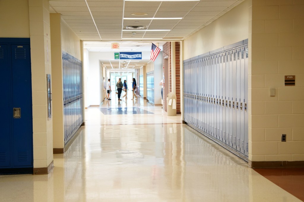 A long high school hallway with blue lockers on both sides, a few students walking in the distance, and an American flag hanging from the wall.
