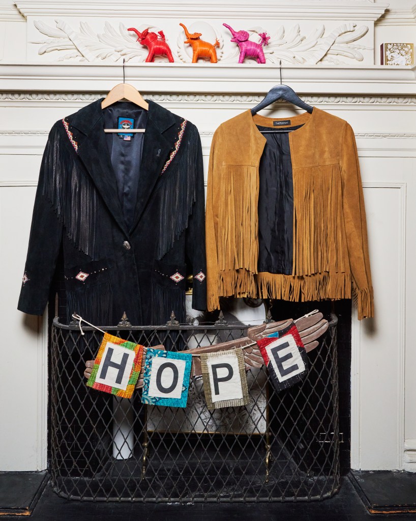Two fringed suede jackets, one black and one brown, hang on a white mantelpiece with small elephant figurines, above a "HOPE" banner.