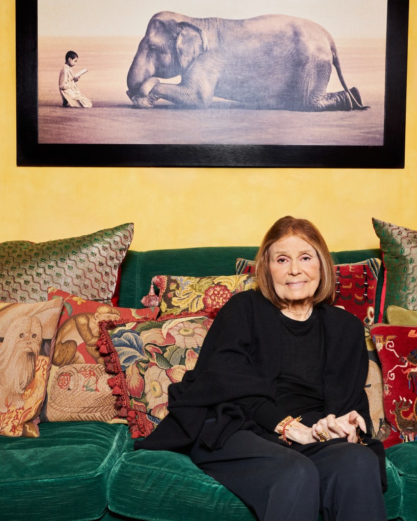 Gloria Steinem seated on a green sofa, surrounded by decorative pillows, with a large framed print of a child reading next to an elephant in the background.