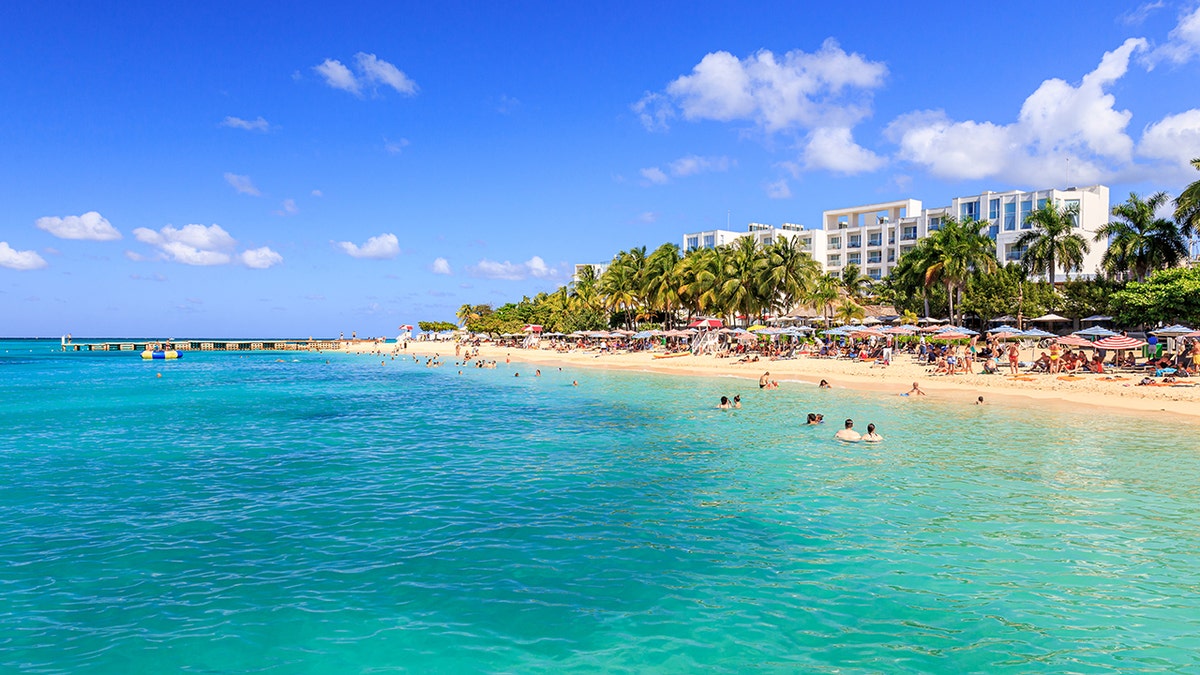 Crowded tropical beach with turquoise water, palm trees, and beachfront resort hotels under a clear blue sky.