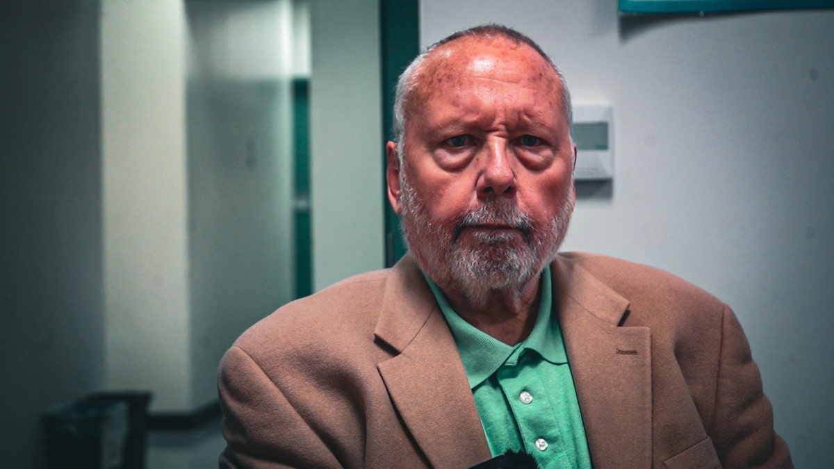 Close up of balding older white man with gray bear in a teal button up shirt and tan sport coat.