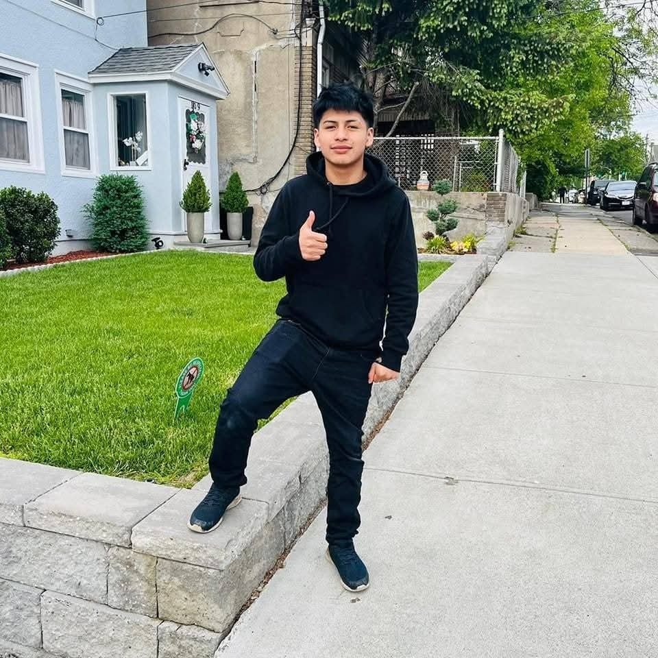 Jonson Temaj, wearing a black hoodie and jeans, gives a thumbs-up while standing on a short stone wall in front of a house.