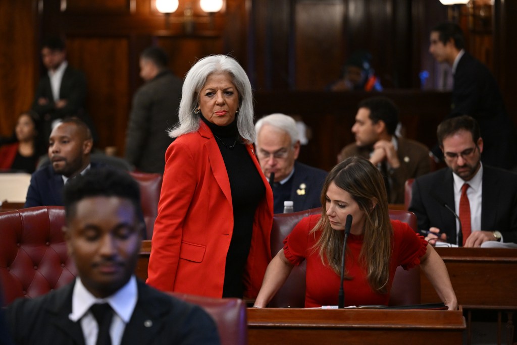 A woman with white hair in a red blazer stands in a room with a woman in a red dress seated next to her, both in a crowd of people.