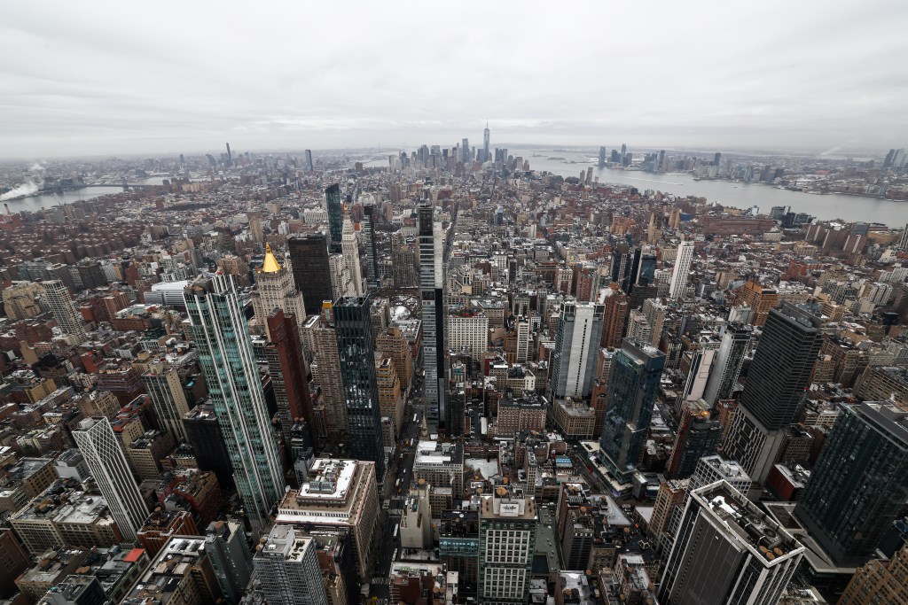 Aerial view of Lower Manhattan skyline on a cloudy day, with numerous skyscrapers and the Hudson River visible in the distance.