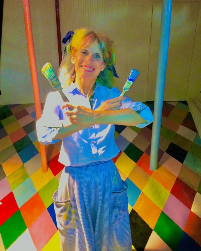 Victoria MacKenzie-Childs holding two paintbrushes with a colorful checkerboard floor.