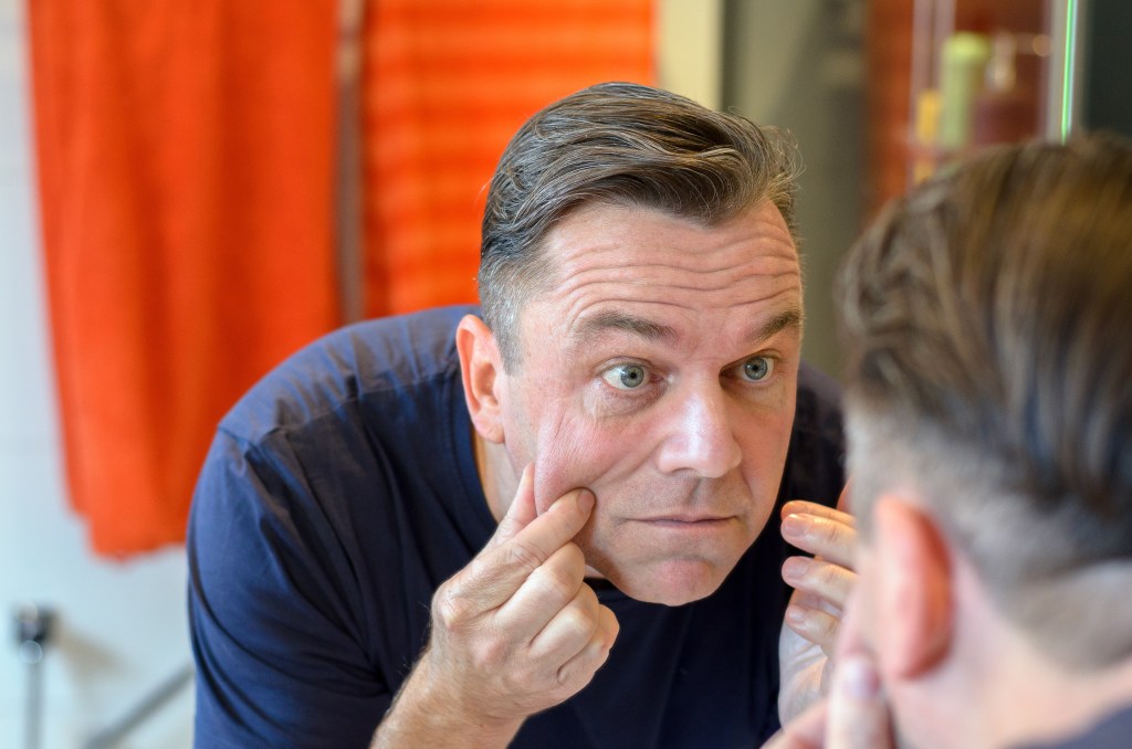 A middle-aged man in a blue shirt looks in a mirror and pinches the skin on his face.
