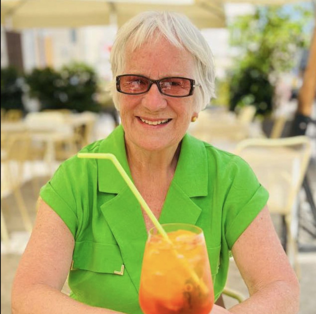 Miriam Lancaster, 84, smiling and wearing a green shirt, holds a drink with a straw.