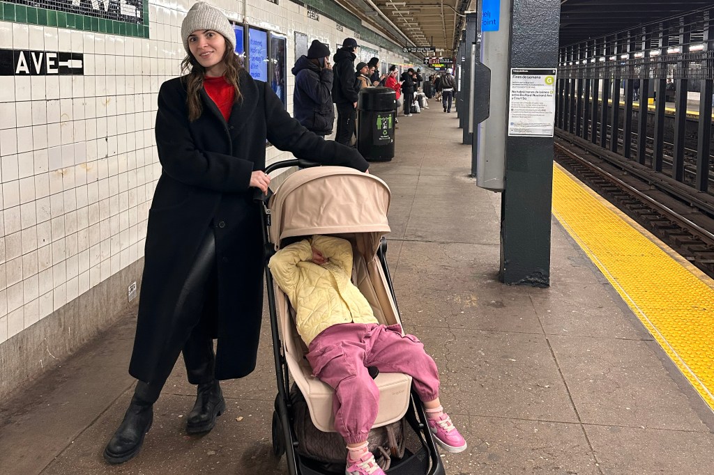 A woman in a black coat and gray beanie pushing a stroller with a child in it on a subway platform.