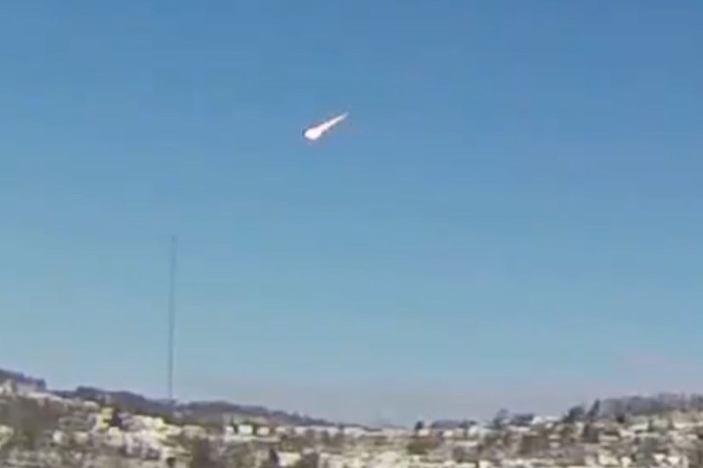 Meteor streaks across a blue sky over a town.