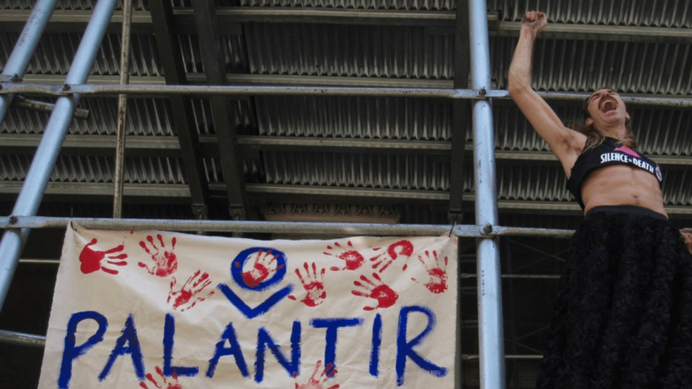 On the scaffolding of a building, a person raises their fist and yells. Beside them, a banner reads "Palantir" with several bloody handprints around it.