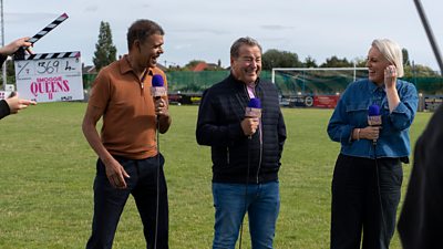 Photo of Chris Kamara, Jeff Stelling and Steph McGovern laughing on a football pitch during filming for Smoggie Queens series two. A clapperboard for the series is held by production crew at the edge of the picture.