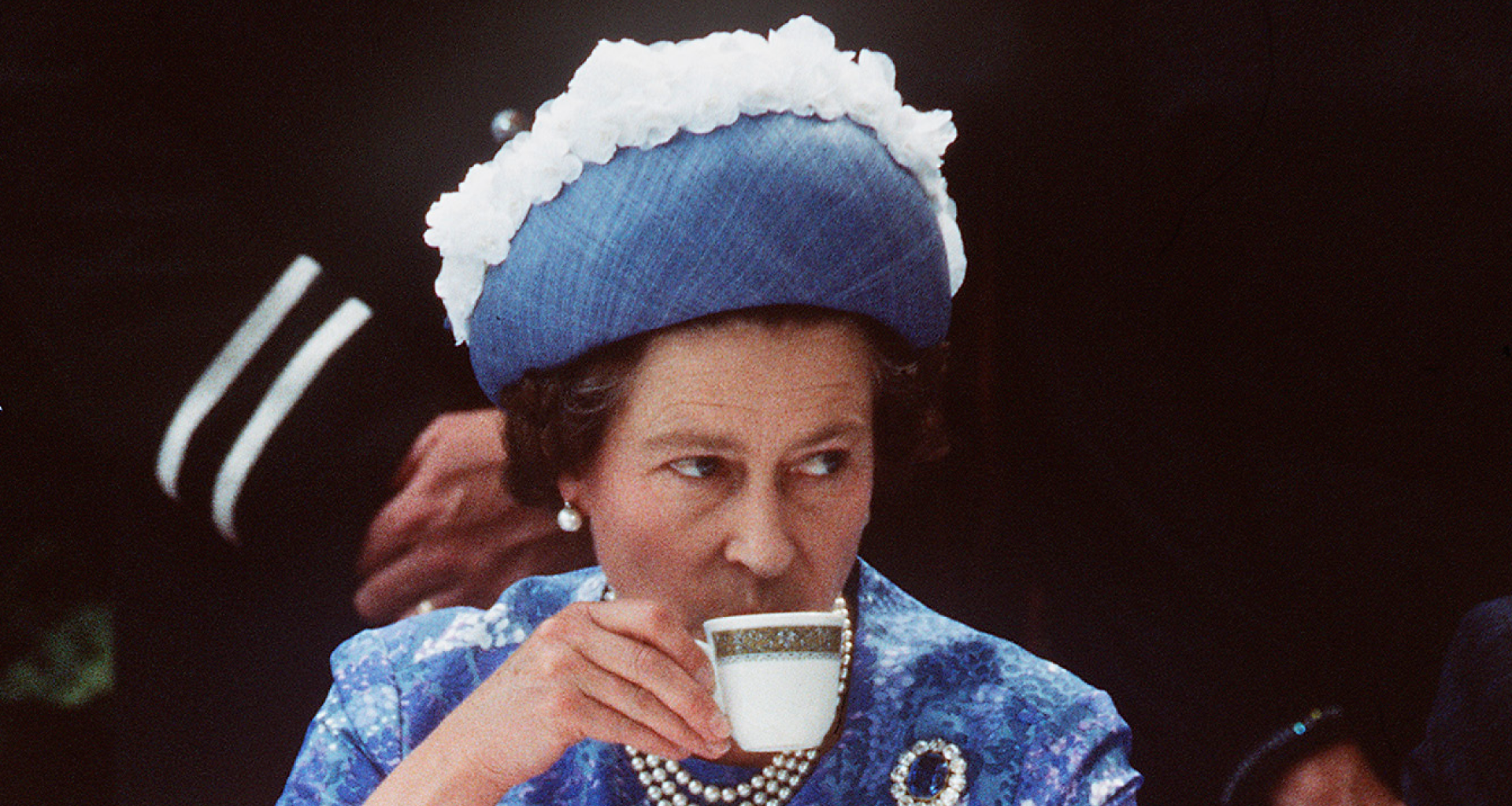 Queen Elizabeth wearing a blue dress and hat drinking tea