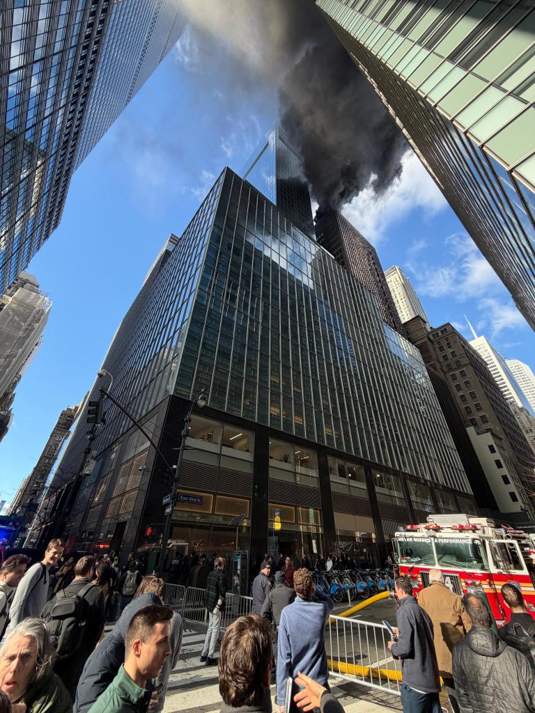 Black smoke rises from a rooftop fire between Madison Ave and 5th Ave in New York City as onlookers and a fire truck are visible on 43rd Street.