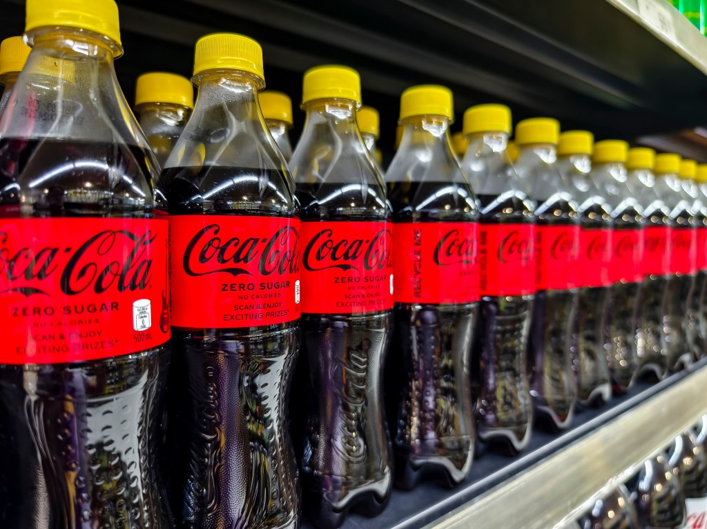 Row of Coca-Cola Zero Sugar bottles with yellow caps on a shelf.