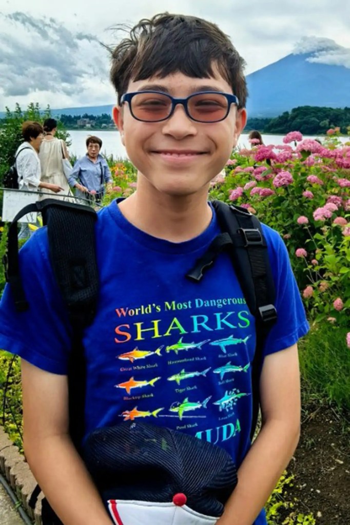 Smiling young man with glasses and a blue "World's Most Dangerous Sharks" T-shirt, carrying a backpack, with a lake and mountain in the background.