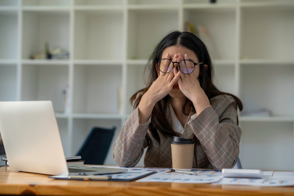 A stressed woman covering her eyes with her hands, sitting at an office desk with a laptop, paper cup, and documents.