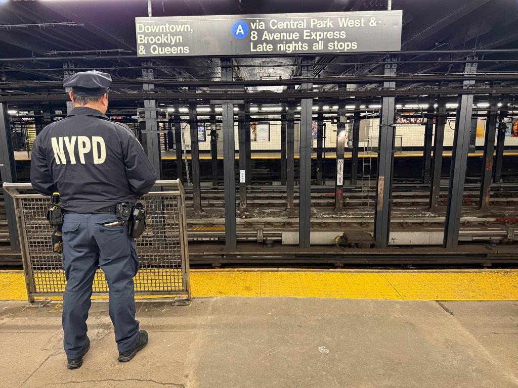 A New York Police Department (NYPD) officer stands on a subway platform looking at the tracks and signs for the A train to Downtown, Brooklyn, and Queens.