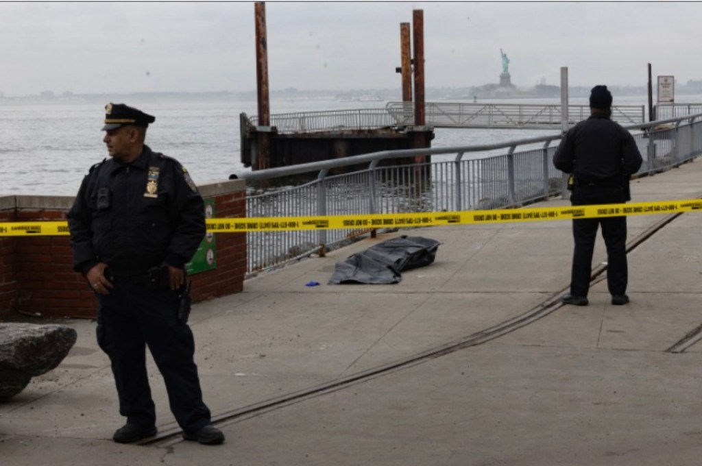 Two police officers stand behind a yellow crime scene tape near a pier with the Statue of Liberty in the background.
