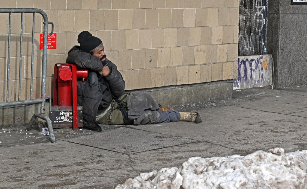 A homeless man sitting on a sidewalk next to a red fire hydrant.