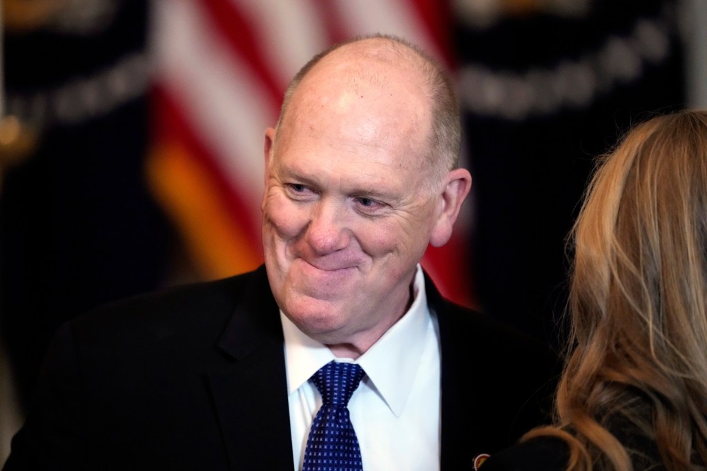 Tom Homan in a black suit and blue patterned tie, smiling with the US flag in the background.