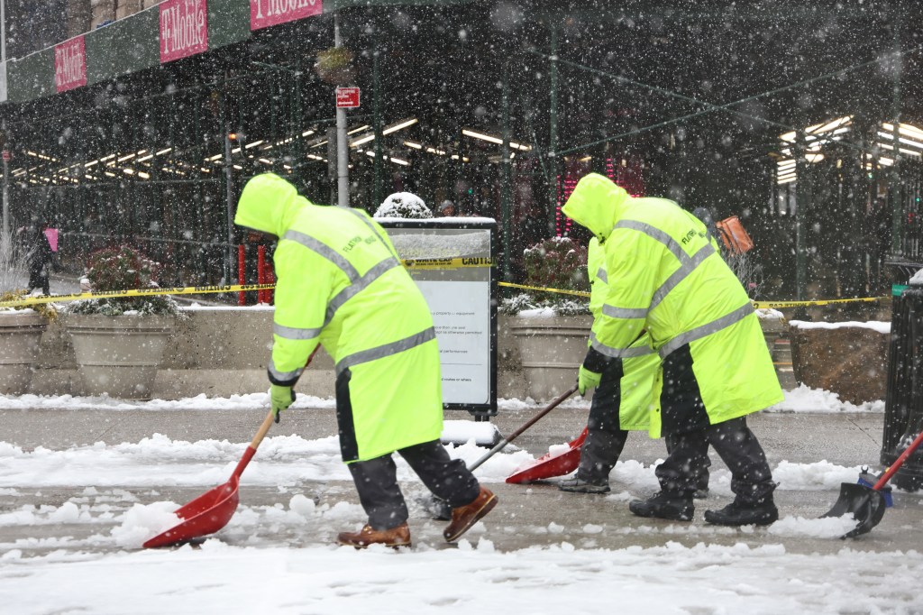 workers in bright yellow work clothes bent over shoveling snow on a city sidewalk