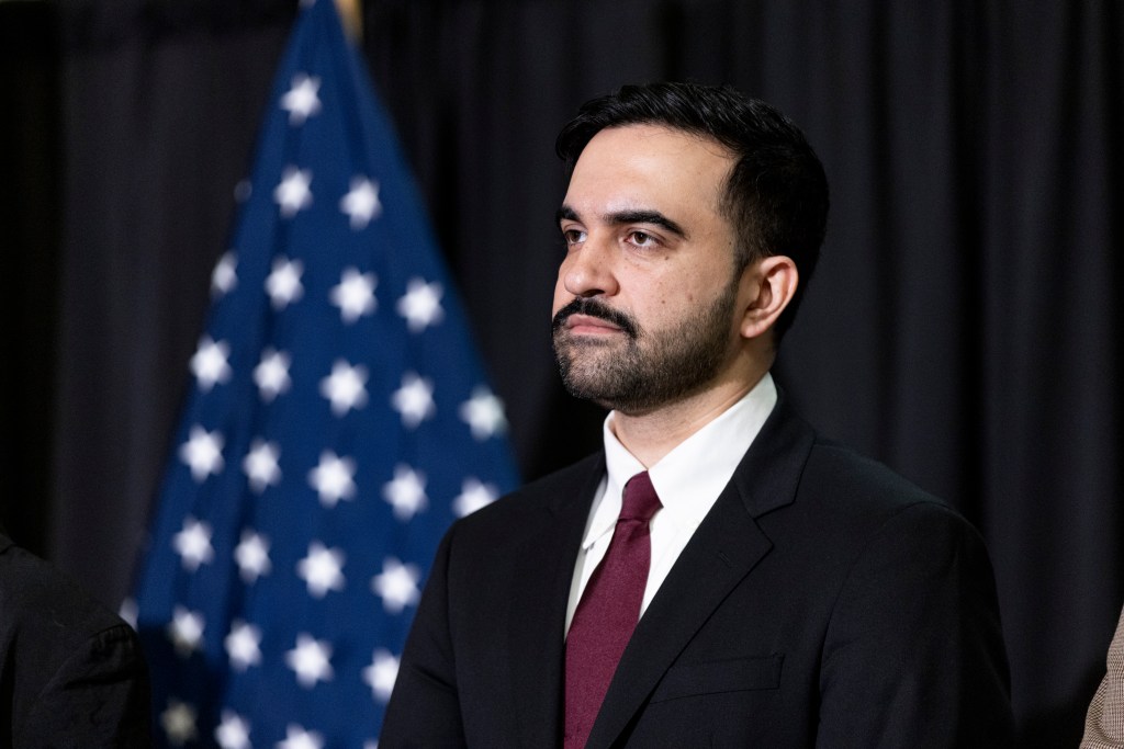 Zohran Mamdani, in a black suit and maroon tie, stands before an American flag.