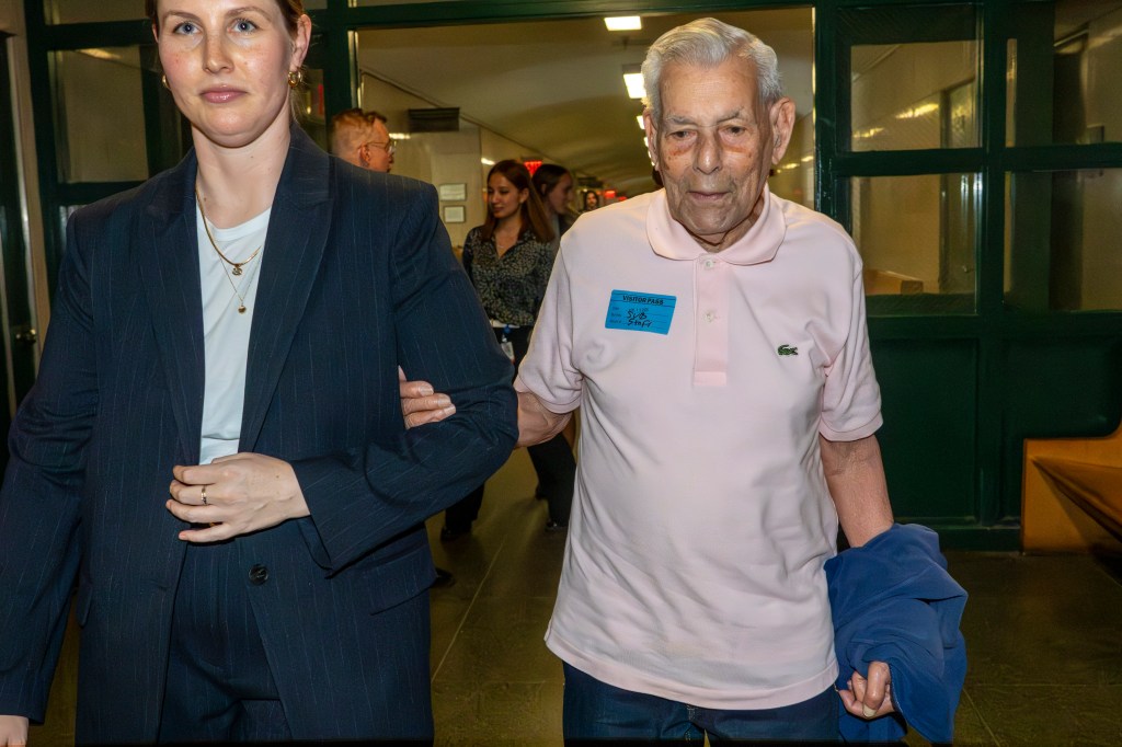A woman in a dark pinstripe suit walks arm-in-arm with an elderly man in a pink polo shirt and a "Visitor Pass" sticker.