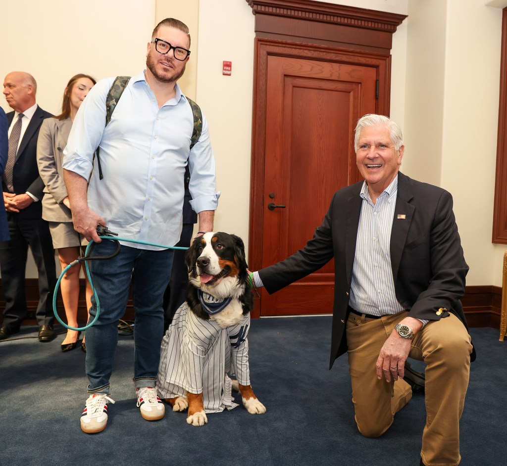 Bruce Blakeman petting a Bernese Mountain Dog wearing a striped shirt and bandana, held on a leash by a man with glasses and a backpack.