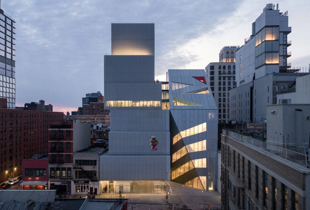 A photo of a contemporary museum building at dusk with a stacked, white geometric facade, featuring a colorful sculpture of a Black adult couple embracing mounted on the exterior, surrounded by city buildings with lit windows.