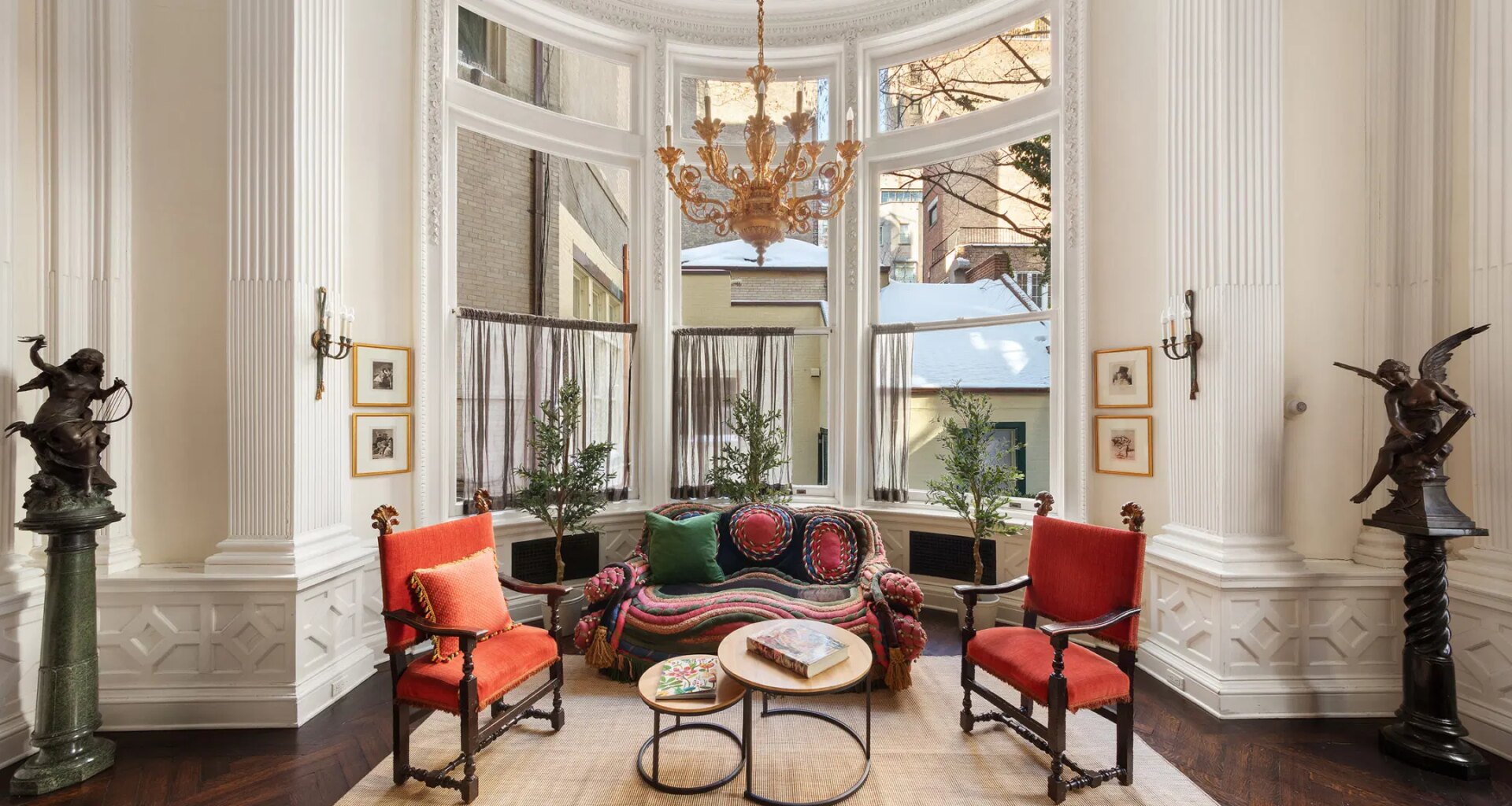 Luxurious sitting room with large bay window, chandelier, ornate decor, red chairs, and view of neighboring buildings.