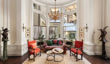 Luxurious sitting room with large bay window, chandelier, ornate decor, red chairs, and view of neighboring buildings.