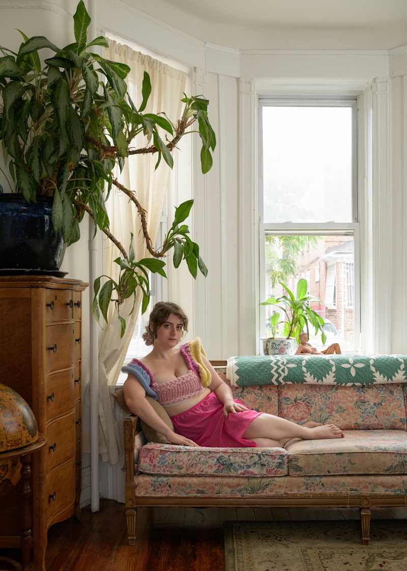 A person lounging on a floral couch in a bright room, wearing a colorful knitted top and pink skirt, with plants and a quilted throw.