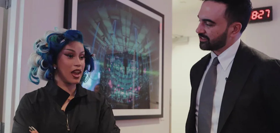 Cardi B, with blue hair rollers, talks to NYC Mayor Zohran Mamdani in a hallway with a digital clock and framed artwork on the wall.