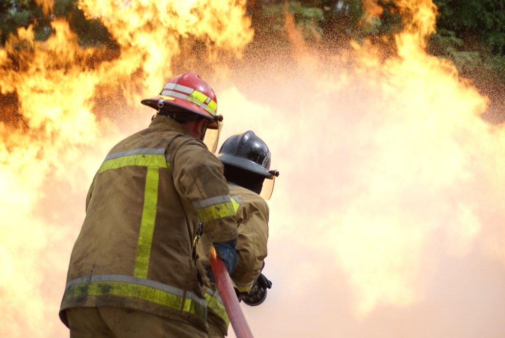 Two firefighters in gear aiming a hose at a large blaze.