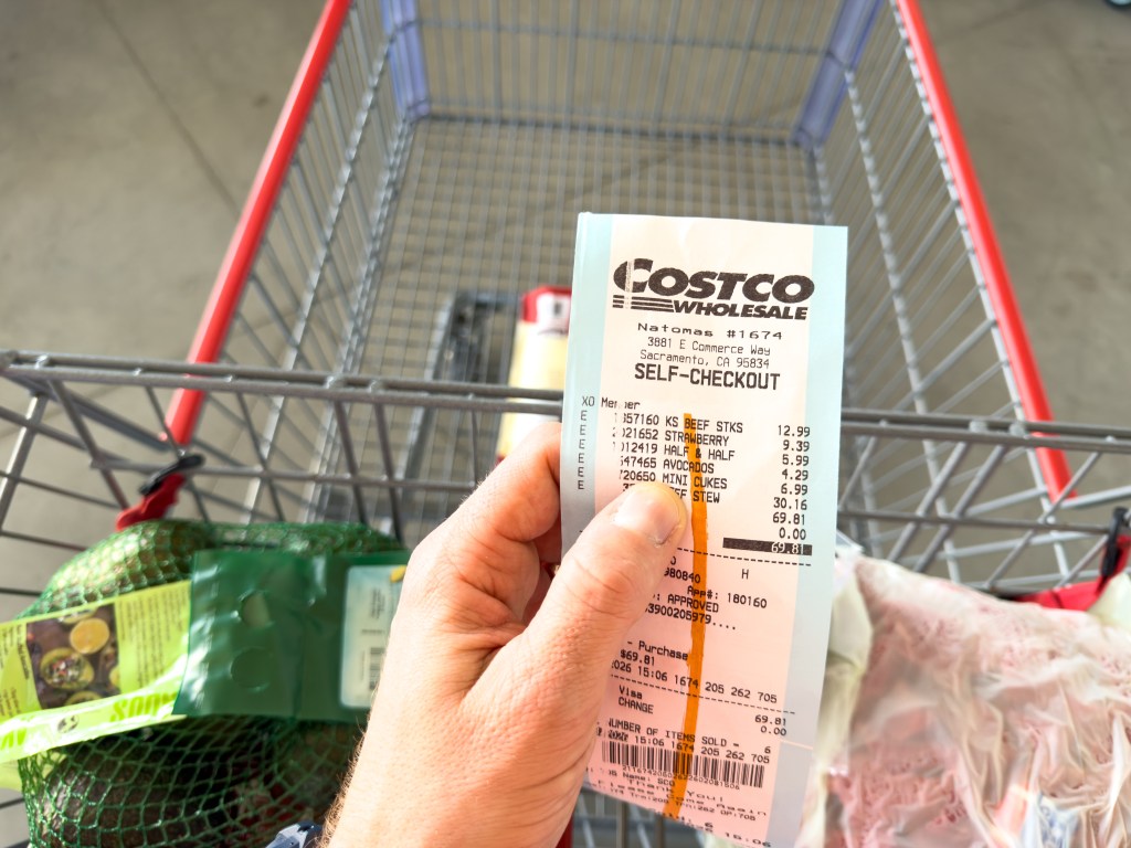 A hand holding a Costco self-checkout receipt over a shopping cart.