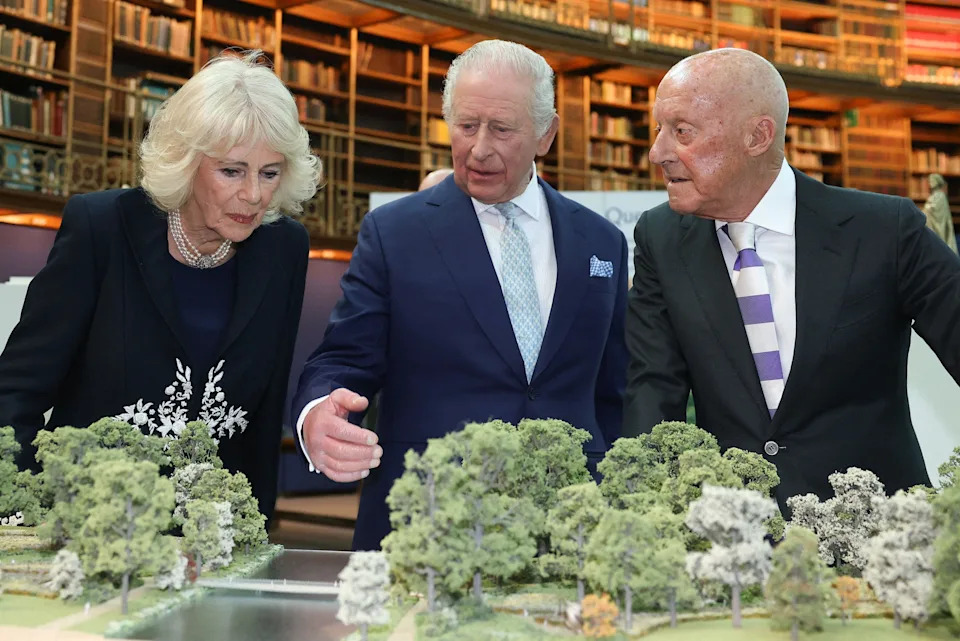 Queen Camilla, King Charles and Norman Foster look at a to-scale model of St James's Park and the design planned for the memorial. They are in the British Museum's library room, so there is a towering wall of books behind them.
