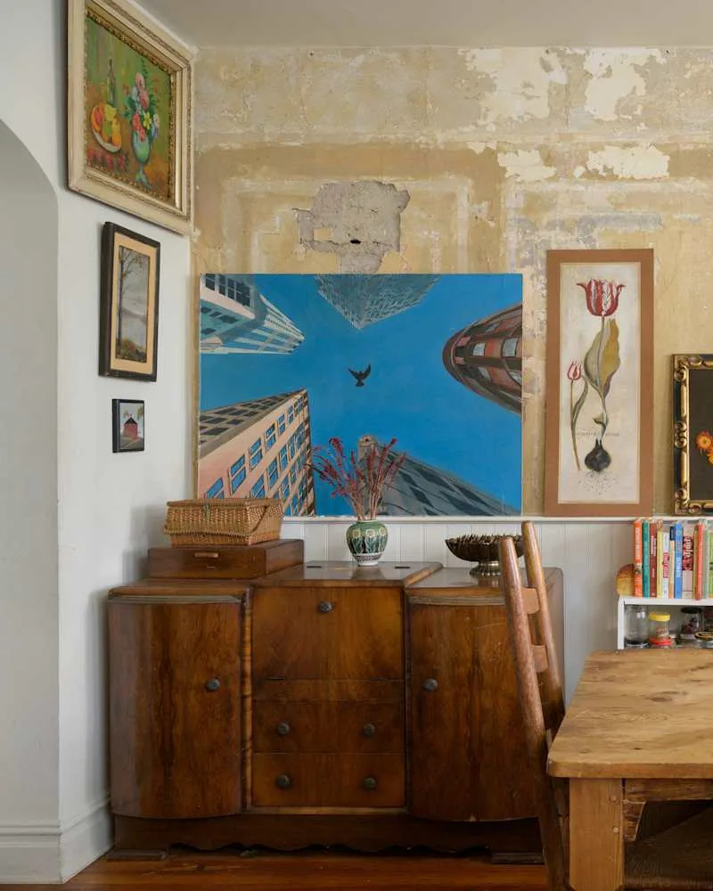 Wooden sideboard with a basket, surrounded by various framed artworks and a dining table with a chair.