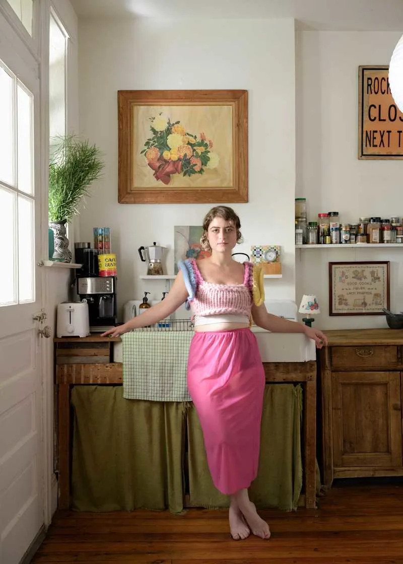 A woman in a pink skirt and colorful knitted top stands in a bright kitchen with wooden accents, plants, and artwork.