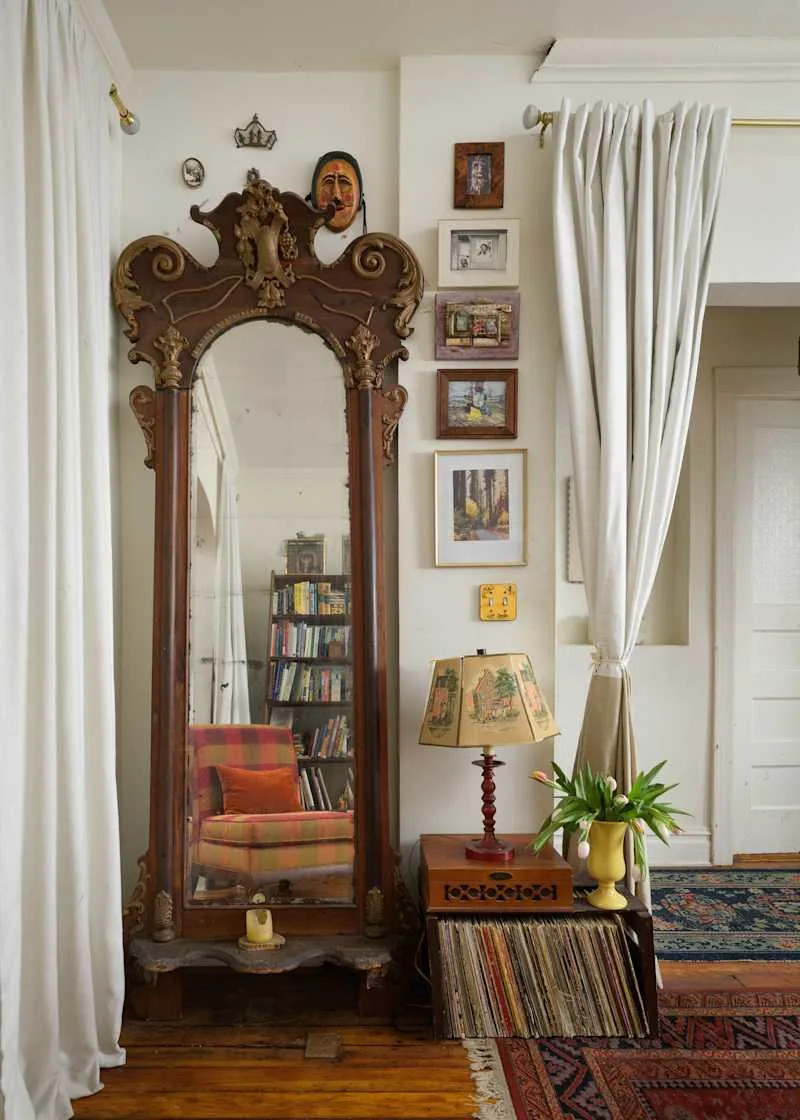 Antique mirror with ornate frame beside a bookshelf, a patterned chair, a lamp with a decorative shade, and a vase of tulips.