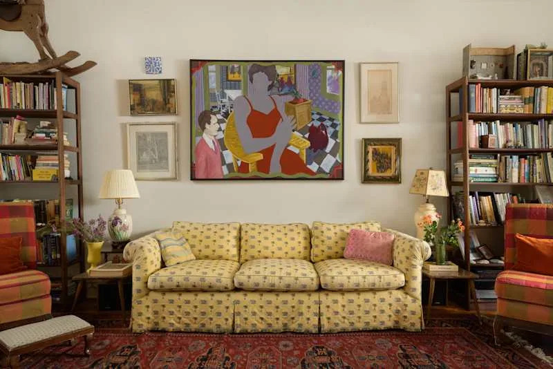Cozy living room with a yellow patterned sofa, colorful artwork, bookshelves, and a floral lamp.