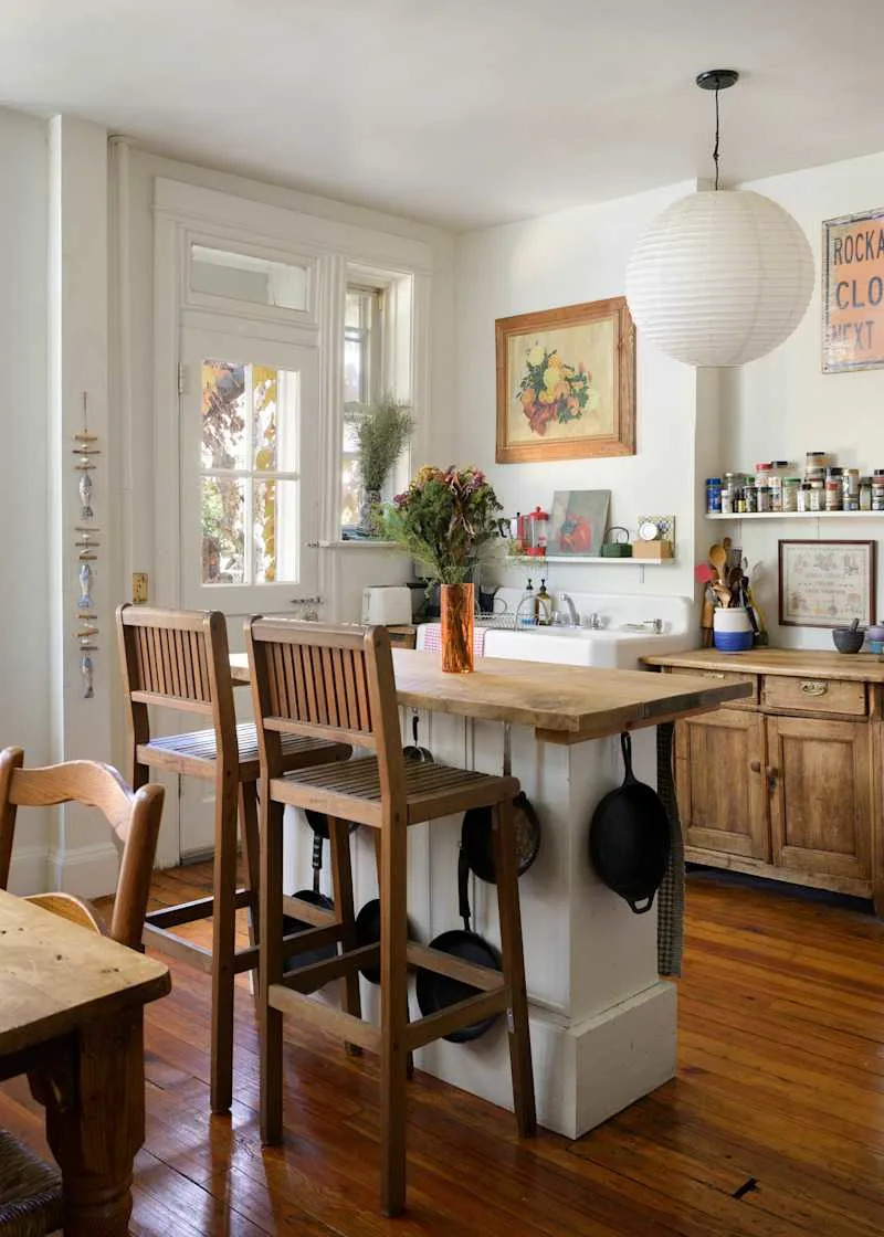 Cozy kitchen with wooden bar stools, a rustic table, potted flowers, and hanging cookware near a window.