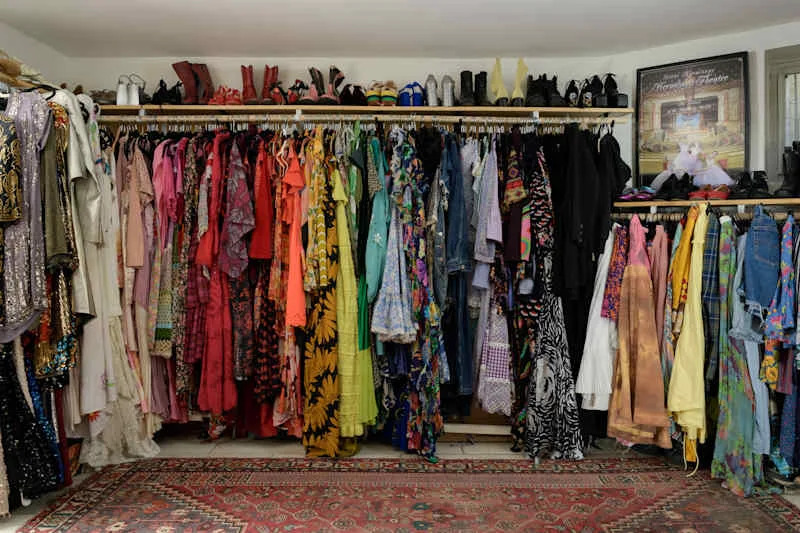 Colorful assortment of dresses and clothing hanging on racks, with various shoes displayed above. A patterned rug is visible below.